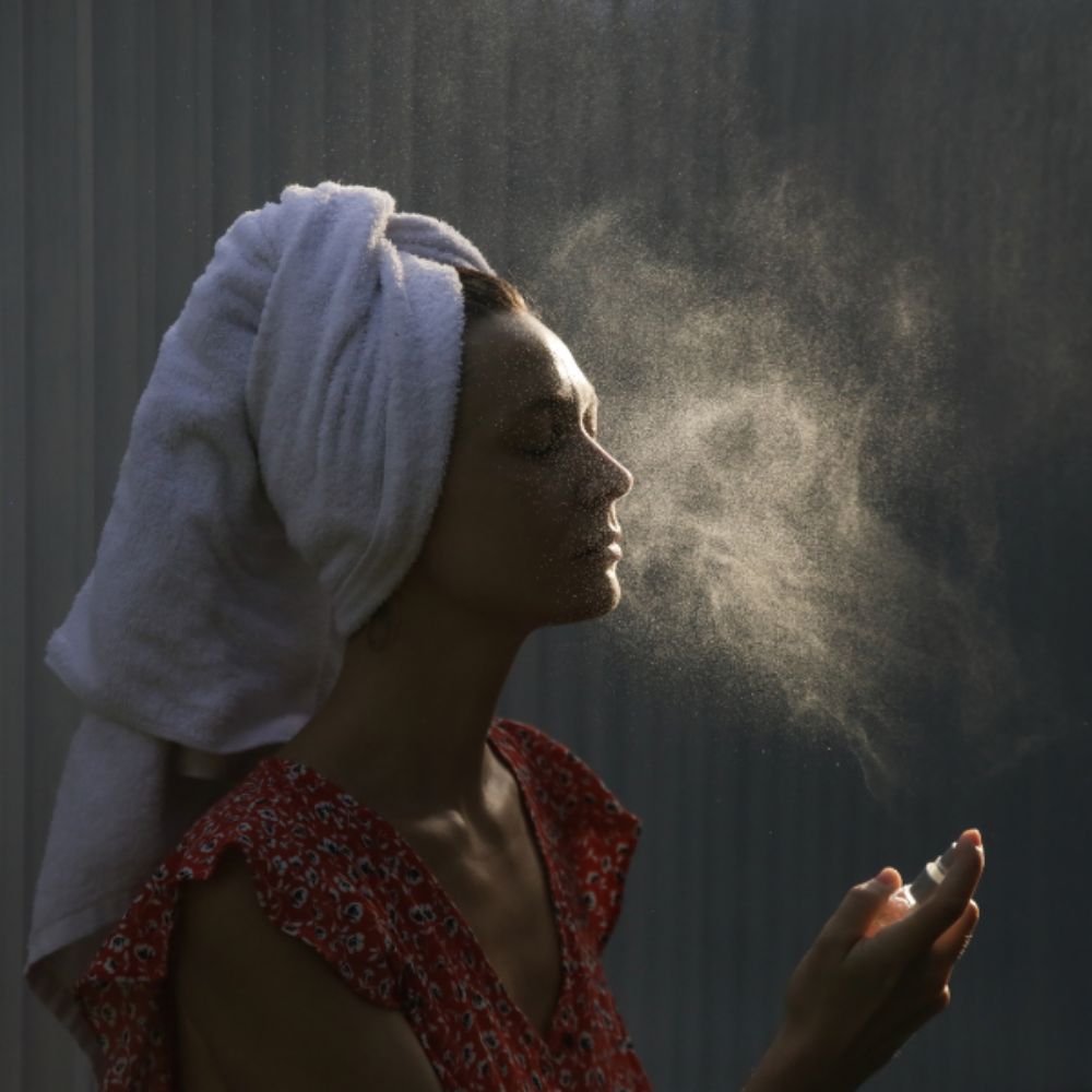 Woman applying mist to her face with a towel on her head against a dark background